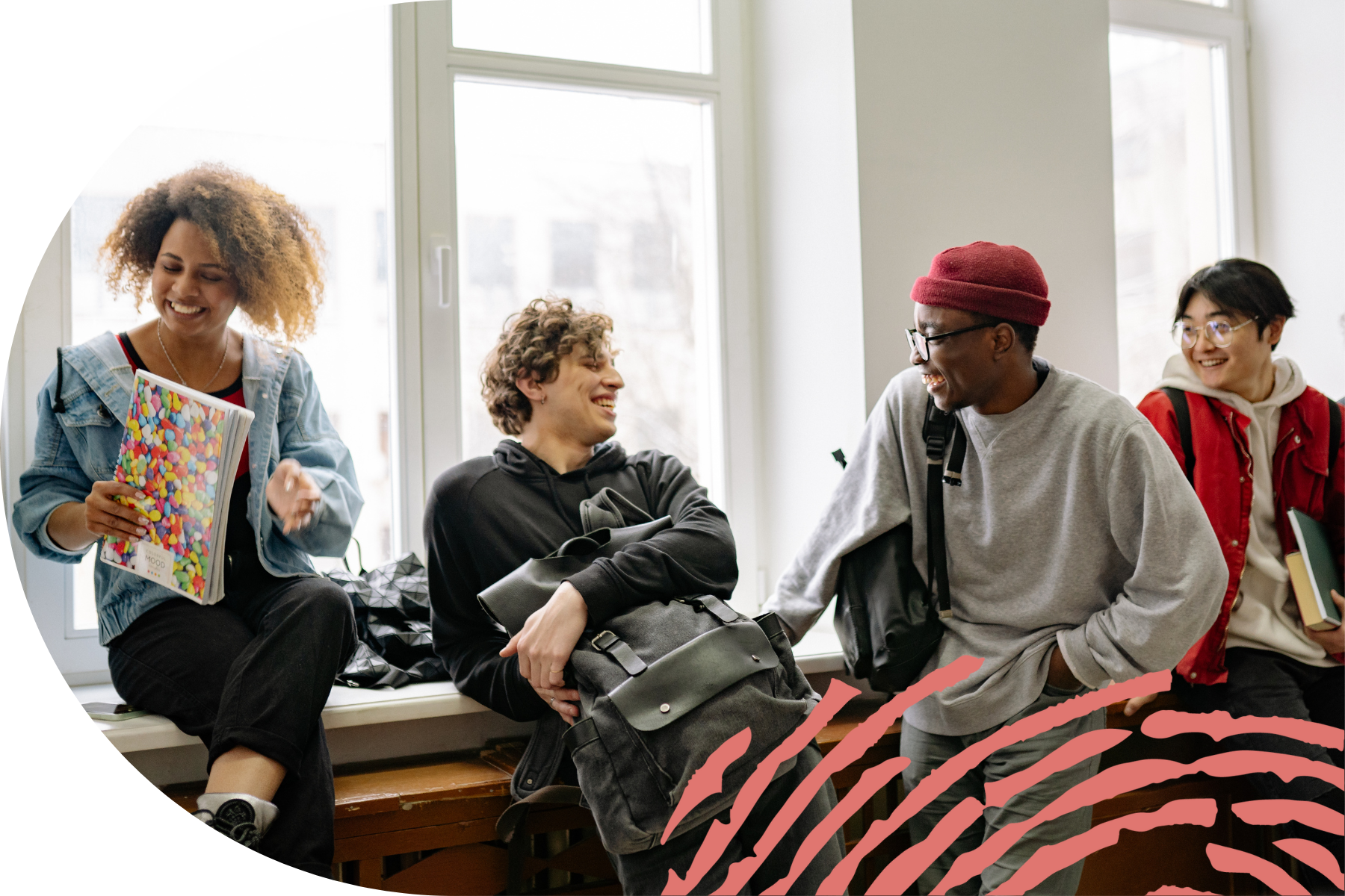 A group of four young adults smiling and chatting while sitting by a large window, holding books and backpacks.